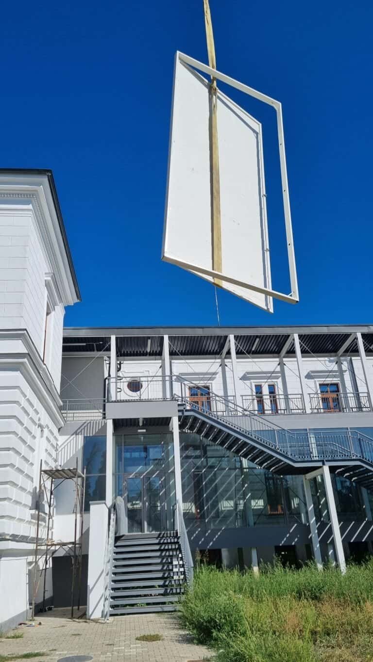 Panou public suspendat în aer, montat pe un suport metalic, în fața unei clădiri moderne, sub cer al.