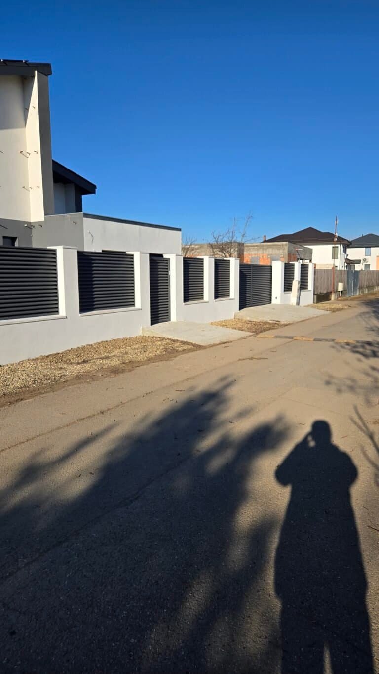 Case moderne cu garduri metalice și ziduri în fața unei străzi liniștite, sub cer senin, cu umbră de.